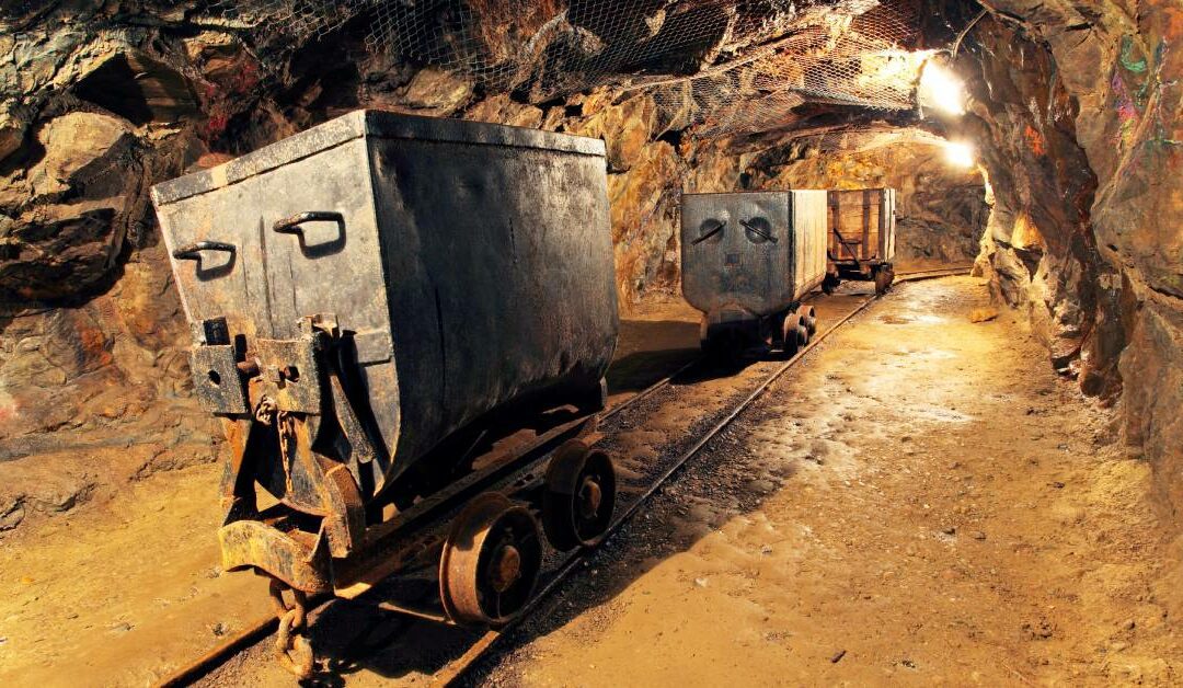 Three metal and wooden rusty mine carts on a track in a cavern with a bright light at the end of it.