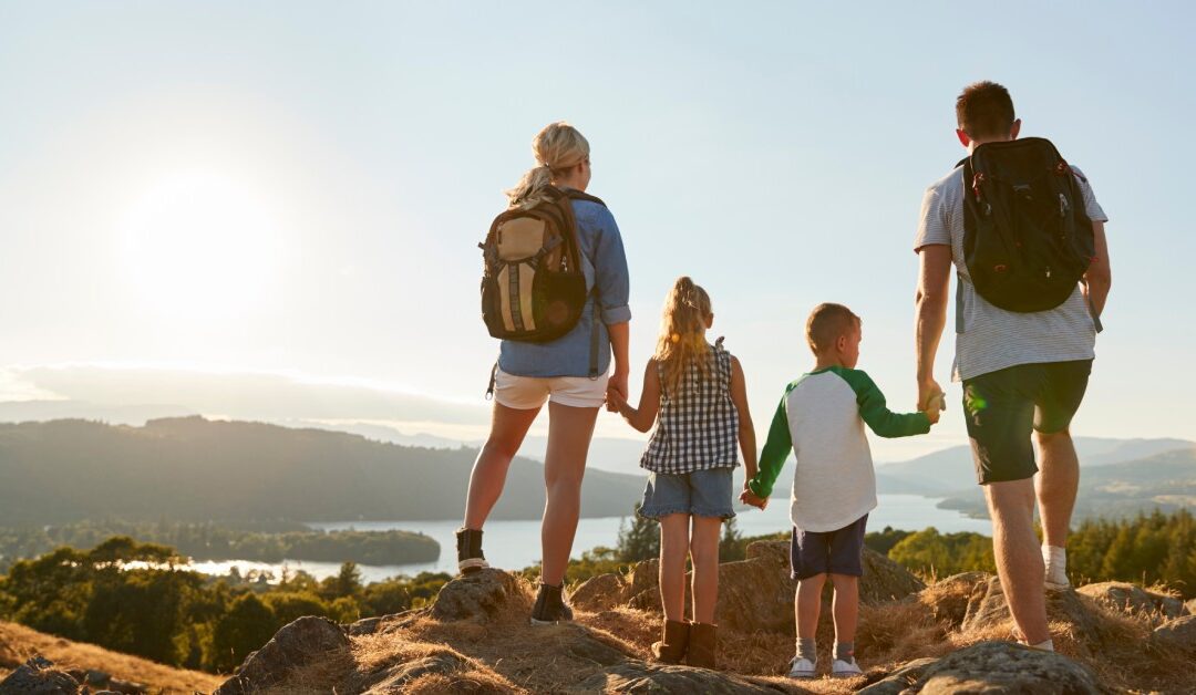 A family of four stands on a mountainside at sunset, looking out over distant mountain peaks and a body of water below.