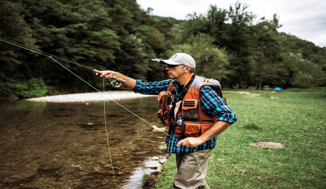 A senior man wearing a baseball cap and orange vest fly fishes by a river, with tall green trees lining the opposite shore.
