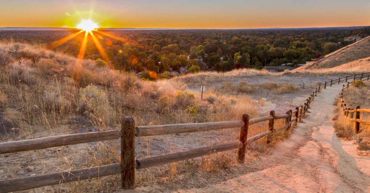 A fenced dirt trail with wooden posts overlooks green foothills and a wide valley glowing under a vivid sunset.