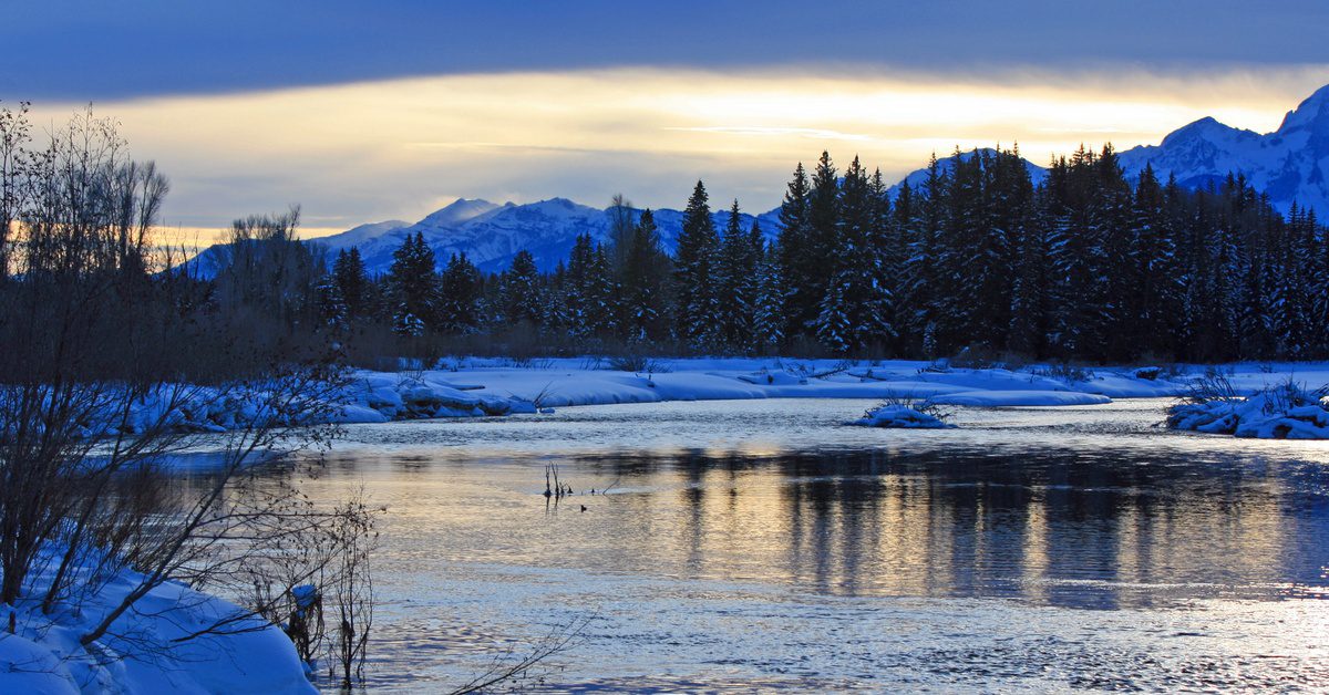 A river flows where water meets fast currents, forming a pool as snow covers trees and mountains at sunset.