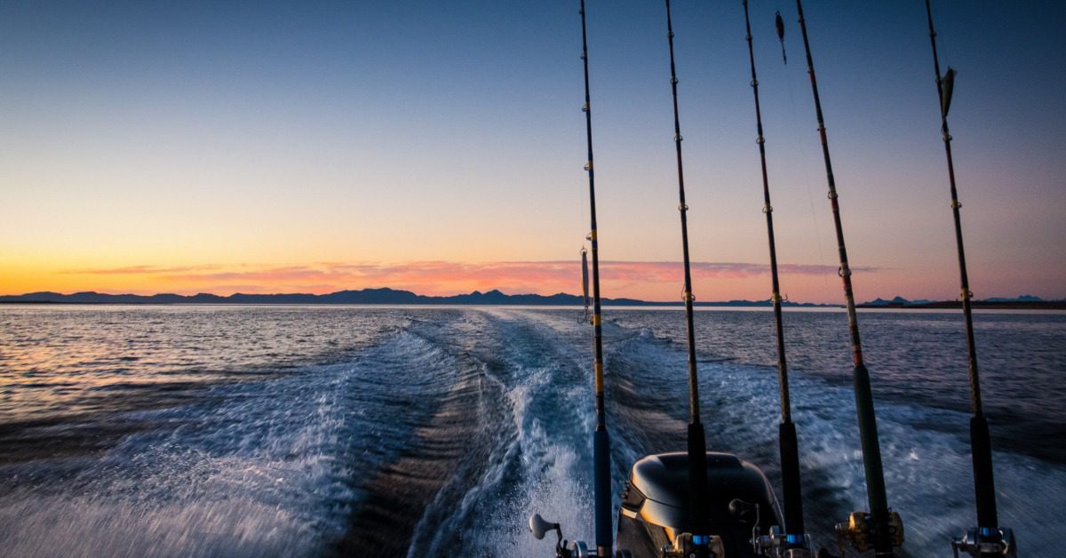 A motorized fishing boat trolls a lake at sunset, with fishing rods lined up along the back of the boat.