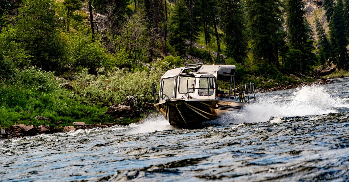 Motorboat speeding through a shallow mountain river with water splashing as it passes forested banks and rocky terrain.