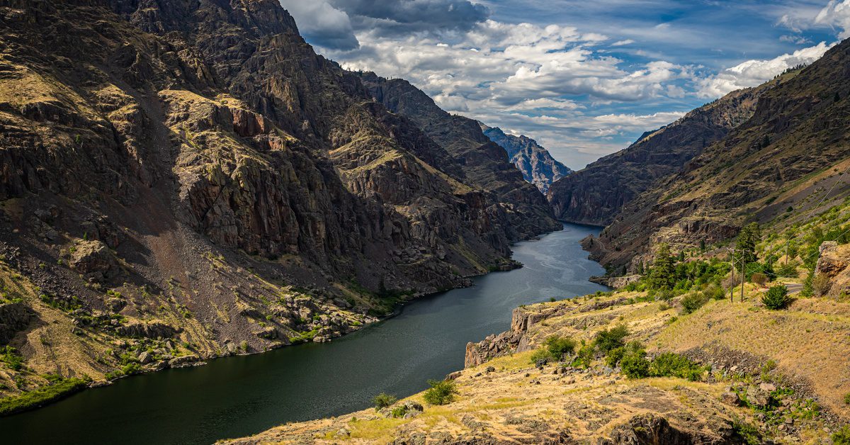 Deep river canyon with steep rocky cliffs, winding waterway, and dramatic clouds stretching across a rugged mountain.