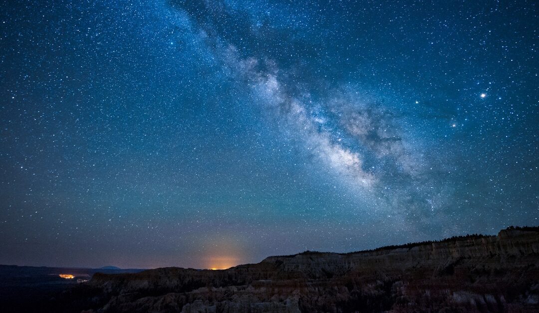 The Milky Way galaxy arches across a star-filled night sky above a rugged canyon landscape at night.