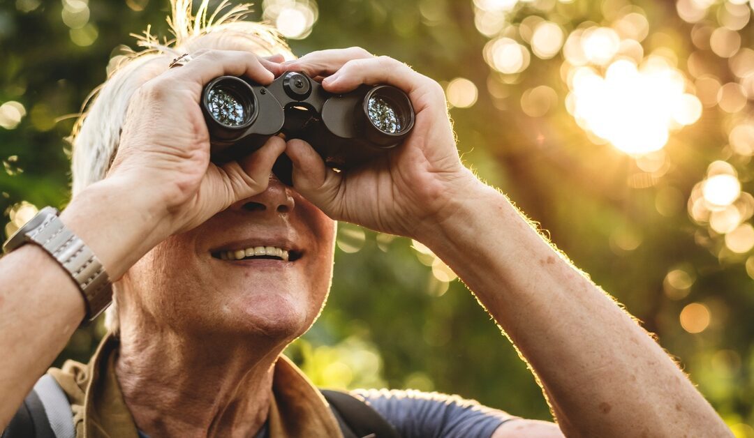 A senior person is smiling while birdwatching with black binoculars outdoors in a sunlit area with trees.